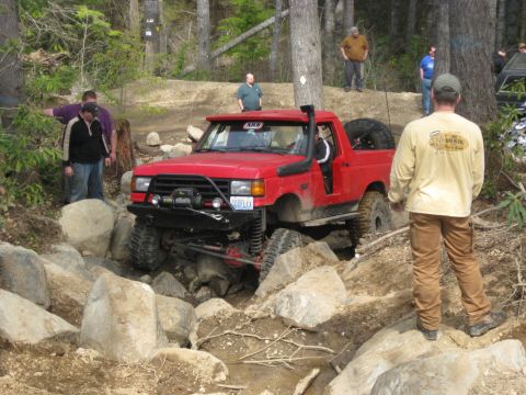 1979 Ford Bronco - A modified red pickup truck tackles a challenging off-road course with spectators observing the action.