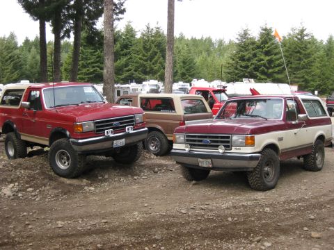 1979 Ford Bronco - Two Ford Broncos, one red and one red and tan, are parked on a gravel lot with trees and RVs in the background.