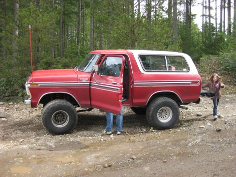 1979 Ford Bronco - Red Ford Bronco with a white top and lifted suspension parked on a dirt road in a wooded area.