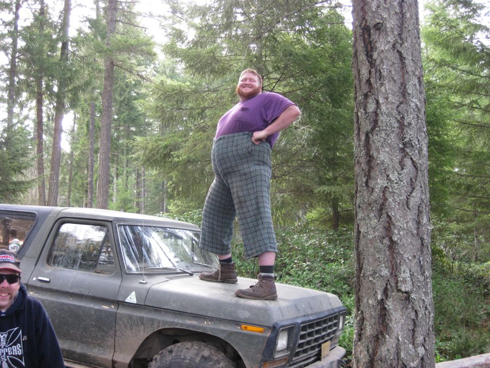 Man in plaid pants stands on the hood of a 1979 Ford Bronco in a wooded area. Another person is visible on the left