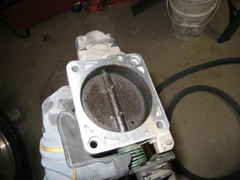 2007 Ford Mustang - A dirty throttle body is held in a gloved hand, showing internal buildup.
