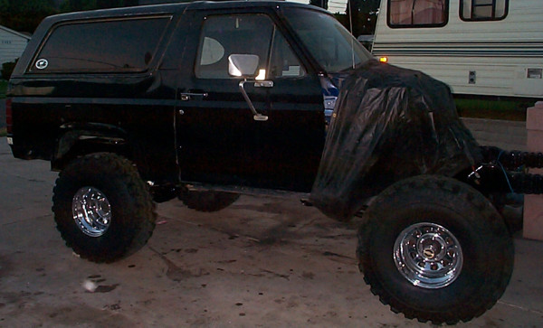 Black two-door Bronco on large mud tires and lift, top off with tarp covering rear, parked by RV at dusk