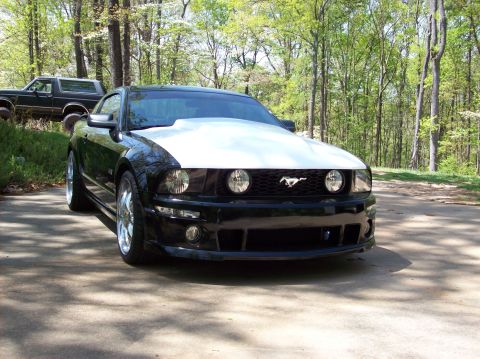 2007 Ford Mustang - A black 2007 Ford Mustang with a prominent white hood and chrome wheels is parked outdoors, with another vehicle visible in the background.