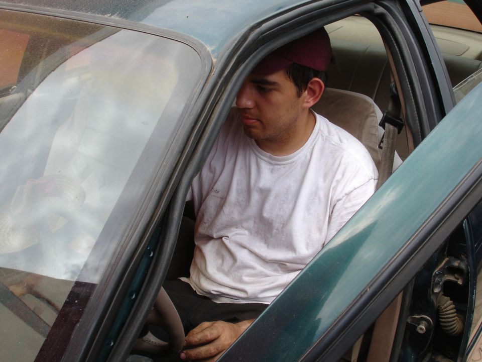 Man wearing a maroon baseball cap sits in the driver's seat of a green car