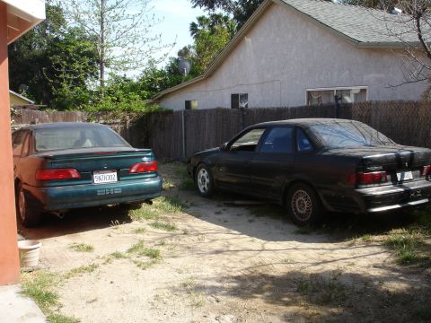 1995 Ford Taurus - Two sedans are parked in a dirt lot with a fence and house in the background.