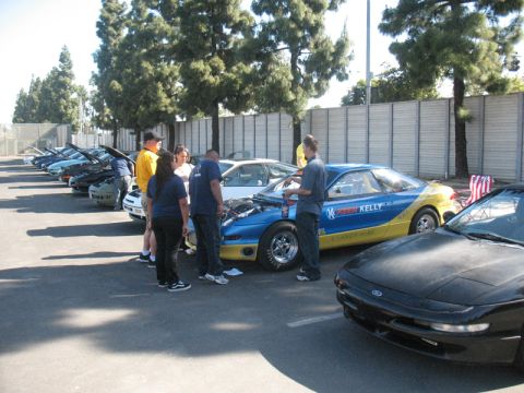 1995 Ford Probe - A group of people gather around a blue and yellow race car with its hood open, parked in a line with other vehicles.