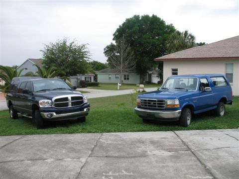 1992 Ford Bronco - A blue 1992 Ford Bronco is parked on the grass next to a dark blue Dodge Ram pickup truck in a residential driveway.