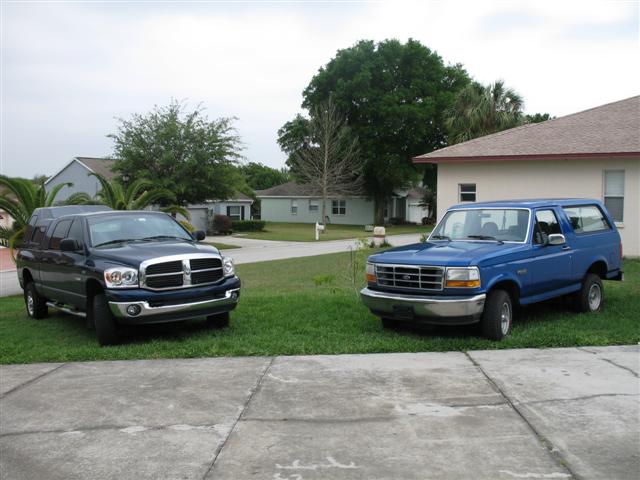 Blue 1990s Ford Bronco parked on grass facing a dark blue Dodge Ram pickup with camper shell