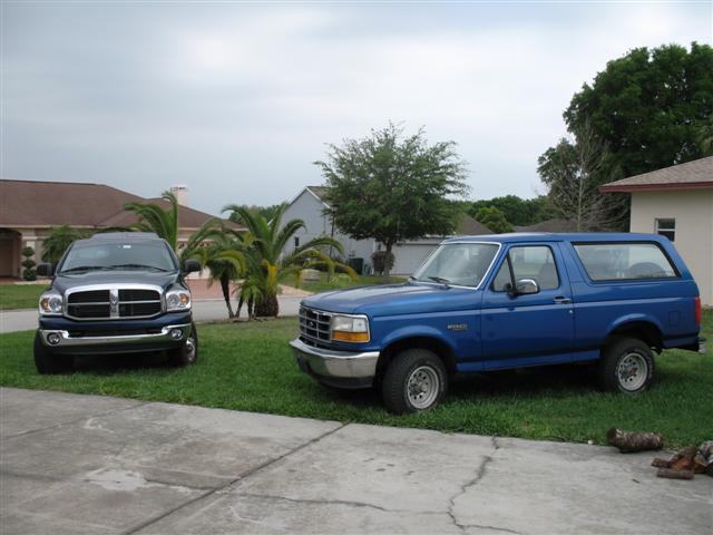 Blue 1990s Ford Bronco parked on grass next to a dark Dodge Ram pickup