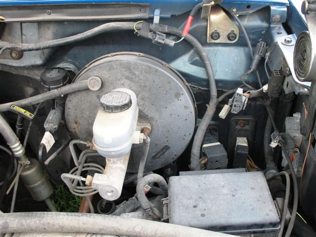 Close-up of a vehicle's engine bay, focusing on the brake master cylinder and booster. Some wiring and a fuse box are visible