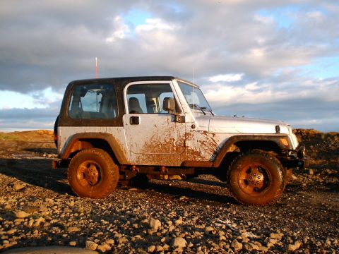 1996 Ford Bronco - A silver Jeep Wrangler, heavily coated in mud, sits on a rocky surface under a cloudy sky.