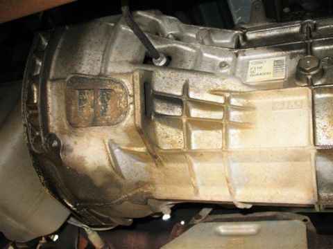 1992 Ford Bronco - Close-up of a cast-aluminum Ford transmission case viewed from underneath, showing the ribbed housing, bellhousing flange, and a Ford oval…