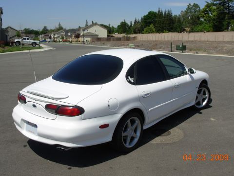 1995 Ford Taurus - This white sedan, likely a 1996 model, is shown from the rear-left quarter on a paved road with houses in the background.