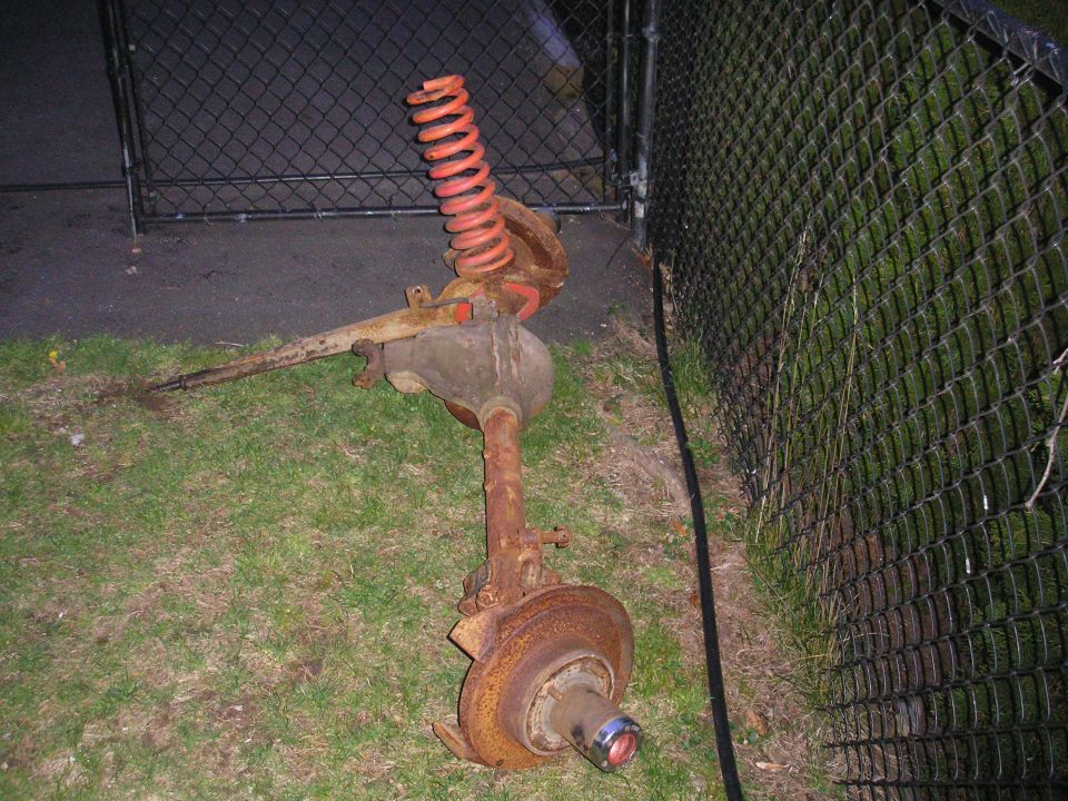 Rusty removed rear axle with differential, drums, and an orange coil spring lying on grass by a fence at night