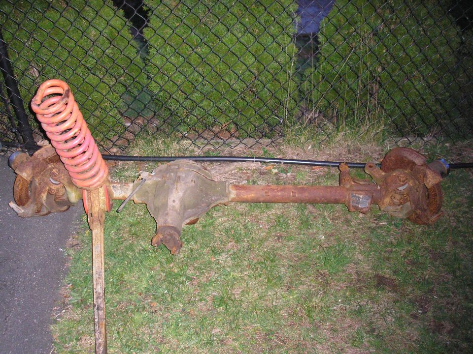 Rusty solid front axle with differential and orange coil spring lying on grass by a chain-link fence