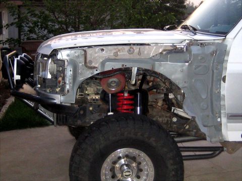 1996 Ford Bronco - Front corner of a white Ford Bronco mid-build with the front fender removed, exposing the inner apron and a red coilover spring over the fr…