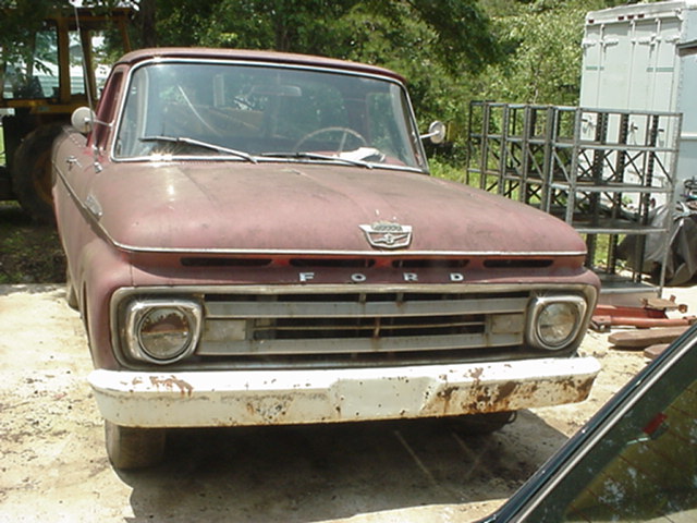 Front view of a rusty, maroon 1961 Ford F-series truck. Grille and bumper are worn