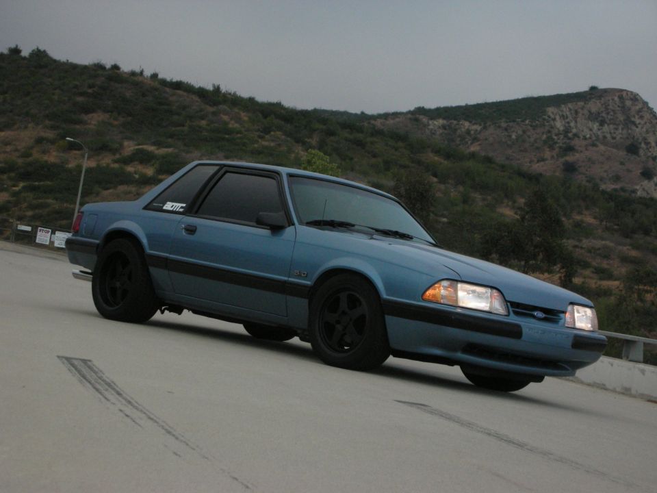 Blue coupe car with black rims on a road with hills in the background. Has a '5.0' badge on the fender