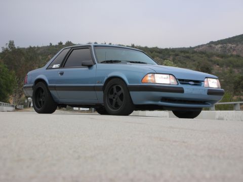 1990 Ford Mustang - A blue 1990 Ford Mustang coupe is parked on a road with hills in the background.