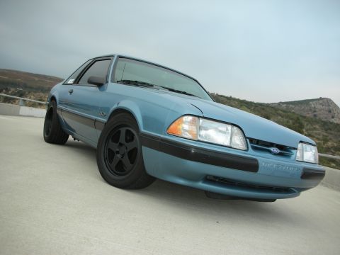 1990 Ford Mustang - A light blue 1990 Ford Mustang is shown from a low angle, highlighting its front quarter view and black aftermarket wheels.