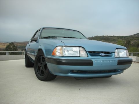 1990 Ford Mustang - This 1990 Ford Mustang features a light blue exterior and aftermarket black wheels, captured from a low front-angle perspective.