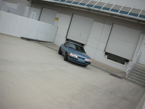 1990 Ford Mustang - This 1990 Ford Mustang coupe is captured in a three-quarter front view, parked in front of industrial loading docks.