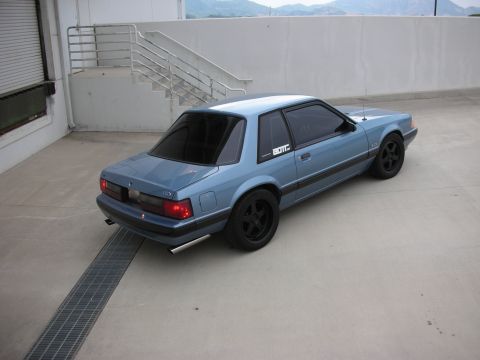 1990 Ford Mustang - A light blue coupe with black accents is viewed from a three-quarter front angle on a concrete surface.