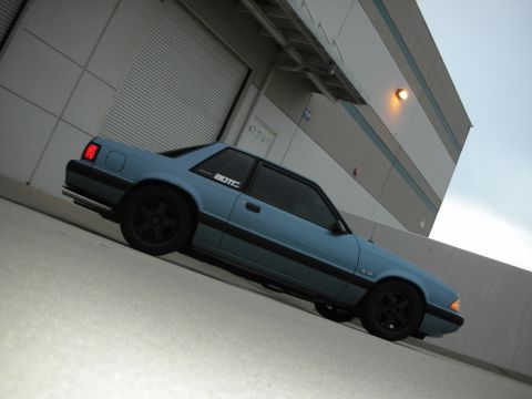 1990 Ford Mustang - 1990 Ford Mustang coupe with black wheels and '5.0' badging parked near a commercial building.
