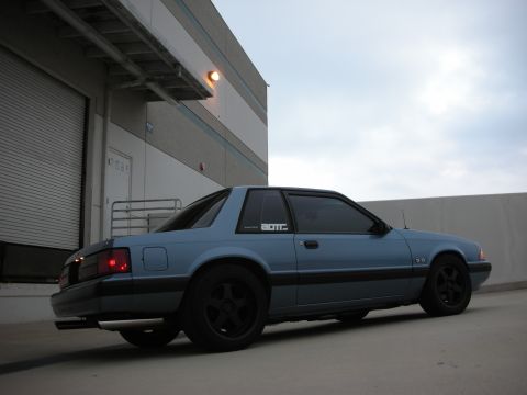 1990 Ford Mustang - A blue 1990 Ford Mustang is parked in front of a commercial building with a cloudy sky overhead.