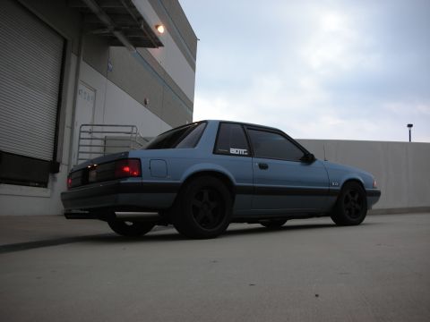 1990 Ford Mustang - A blue 1990 Ford Mustang is viewed from a low angle, showing its side and rear profile against a cloudy sky.