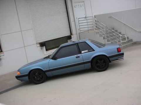 1990 Ford Mustang - A blue 1990 Ford Mustang coupe with black wheels and a "5.0" badge is parked outside a commercial building.
