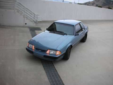 1990 Ford Mustang - A blue coupe is parked on a concrete surface with a metal grate in front of it.