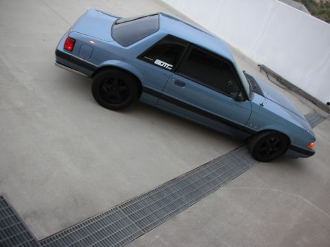 1990 Ford Mustang - A blue coupe with black wheels is parked on a concrete surface with metal grates.
