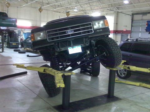 1990 Ford Bronco - A black Ford F-Series truck is raised on a lift inside a garage, showing its suspension and undercarriage.