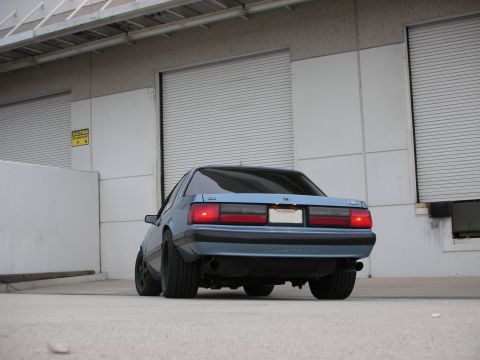 1990 Ford Mustang - Rear view of a blue 1990 Ford Mustang parked in front of a warehouse with closed garage doors.