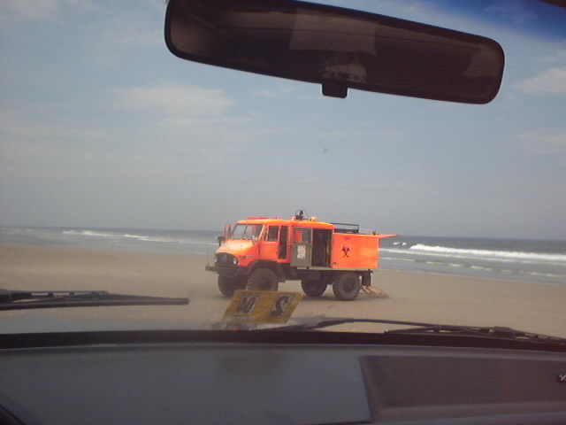Orange off-road truck on a beach seen through a car windshield with rearview mirror visible.