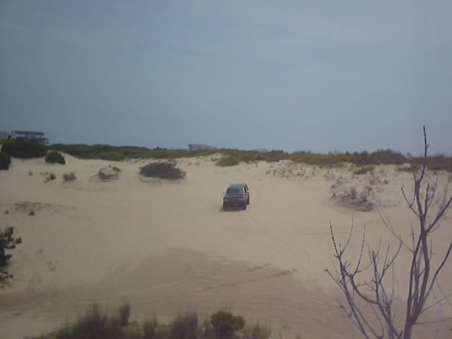 Dark SUV seen from behind driving over sand dunes with brush and distant beach houses