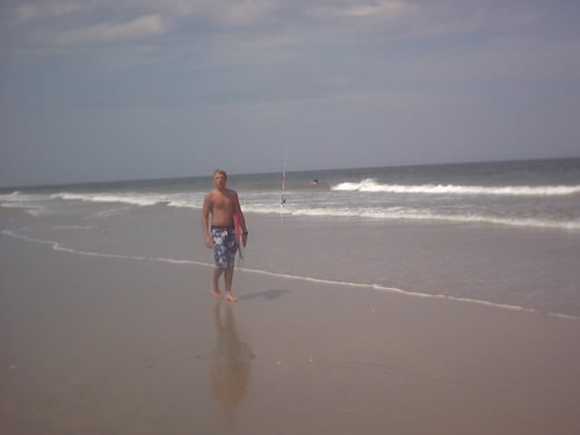 Shirtless man in floral board shorts standing barefoot at the water's edge on a beach with fishing rod behind
