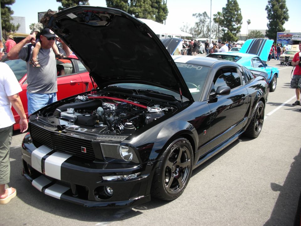 Black Ford Mustang with hood open, showing engine. Another blue car is behind it