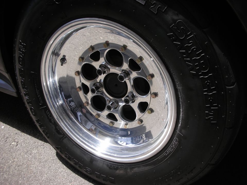 Close-up of a shiny chrome drag racing wheel with a black tire. The tire has text on the sidewall