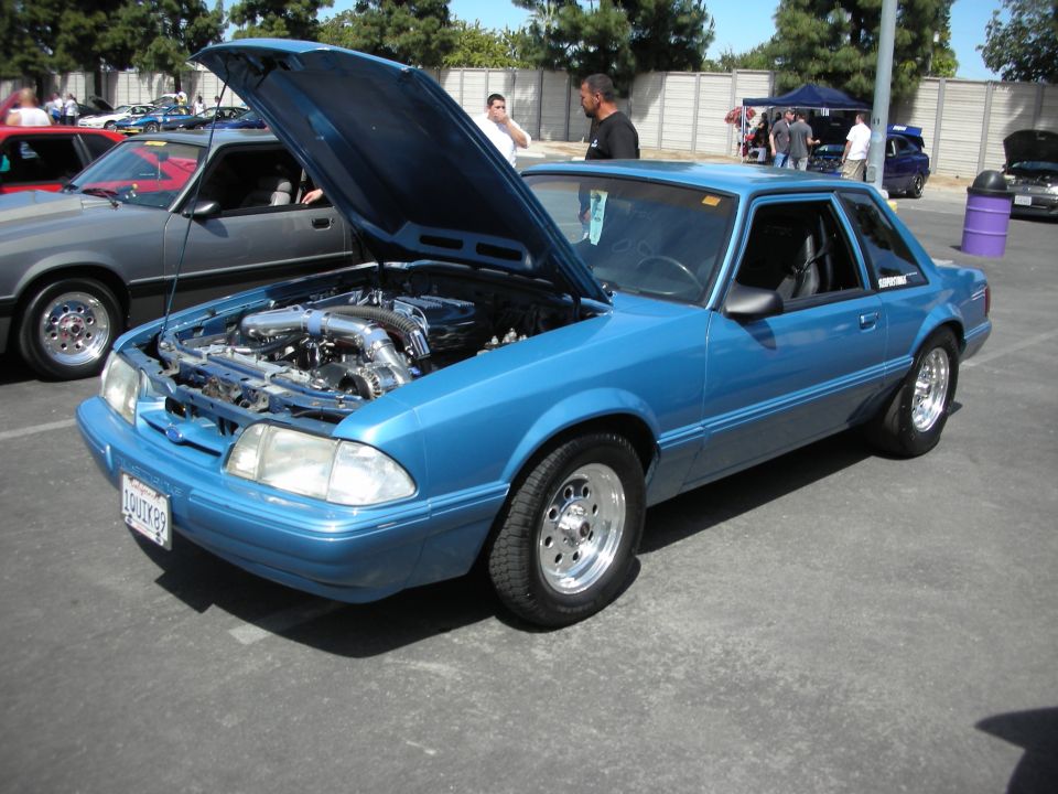 Blue Ford Mustang coupe with aftermarket wheels and engine modifications, hood open. Another car is parked to its left