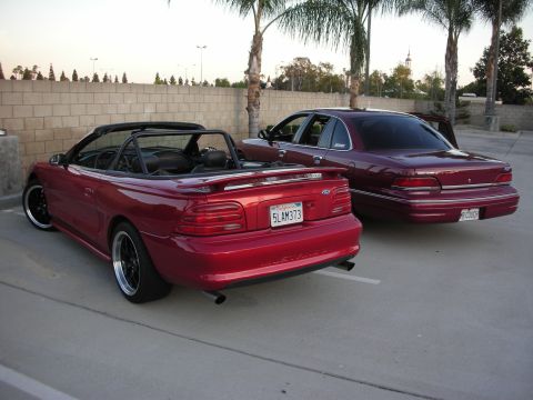 1992 Ford Crown Victoria - A maroon 1992 Ford Crown Victoria sedan is parked next to a red convertible, both seen from the rear.
