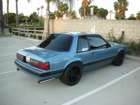 1992 Ford Crown Victoria - This blue 1987 Ford Mustang coupe is parked in a lot with palm trees in the background.
