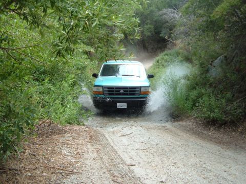 1988 Ford Bronco - Head-on view of a teal Ford pickup blasting through a water crossing on a brush-lined dirt trail, throwing a heavy sheet of spray off the p…