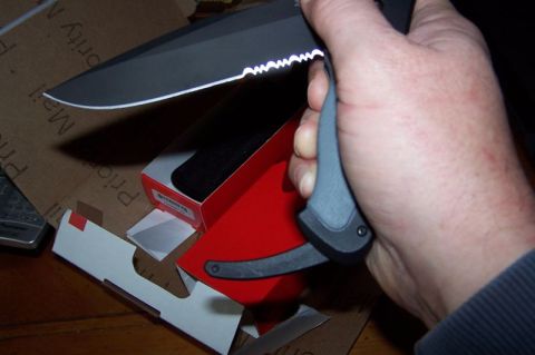 1990 Ford Bronco - A hand grips a black serrated knife, with packaging and a red box visible in the background.