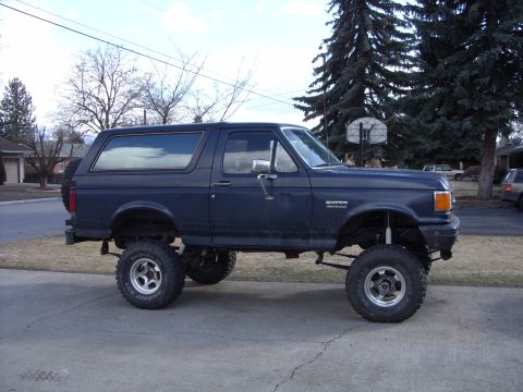 1988 Ford Bronco - Passenger-side profile of a lifted dark blue full-size Ford Bronco with faded paint parked on a suburban driveway with bare trees and a bas…