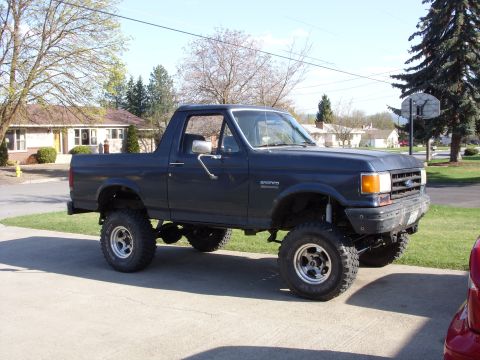 1988 Ford Bronco - Passenger-side profile of a dark blue full-size Ford Bronco with the rear top removed, sitting on a suburban concrete driveway. It runs a t…