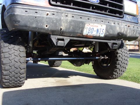 1988 Ford Bronco - Ground-level view under the front of a lifted Ford truck on a concrete driveway, showing the front axle, differential, coil-spring radius a…