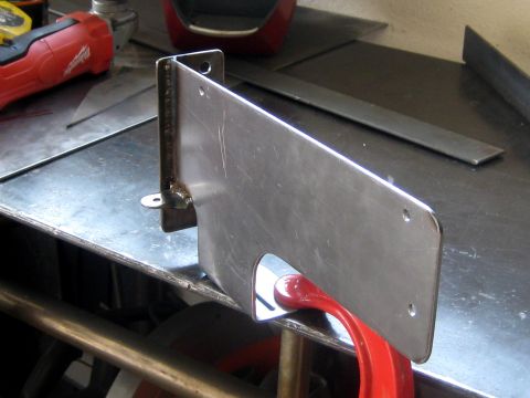 1996 Ford Bronco - A shop-fabricated bare steel license plate bracket clamped to a metal workbench, with a welded angle-iron upright, a small tab foot and dri…