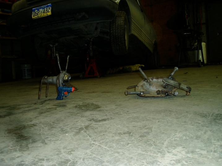 Two scrap metal robot sculptures on a garage floor with a car raised on jack stands.